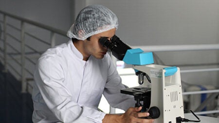 Man with white coat and hair net looking into a microscope