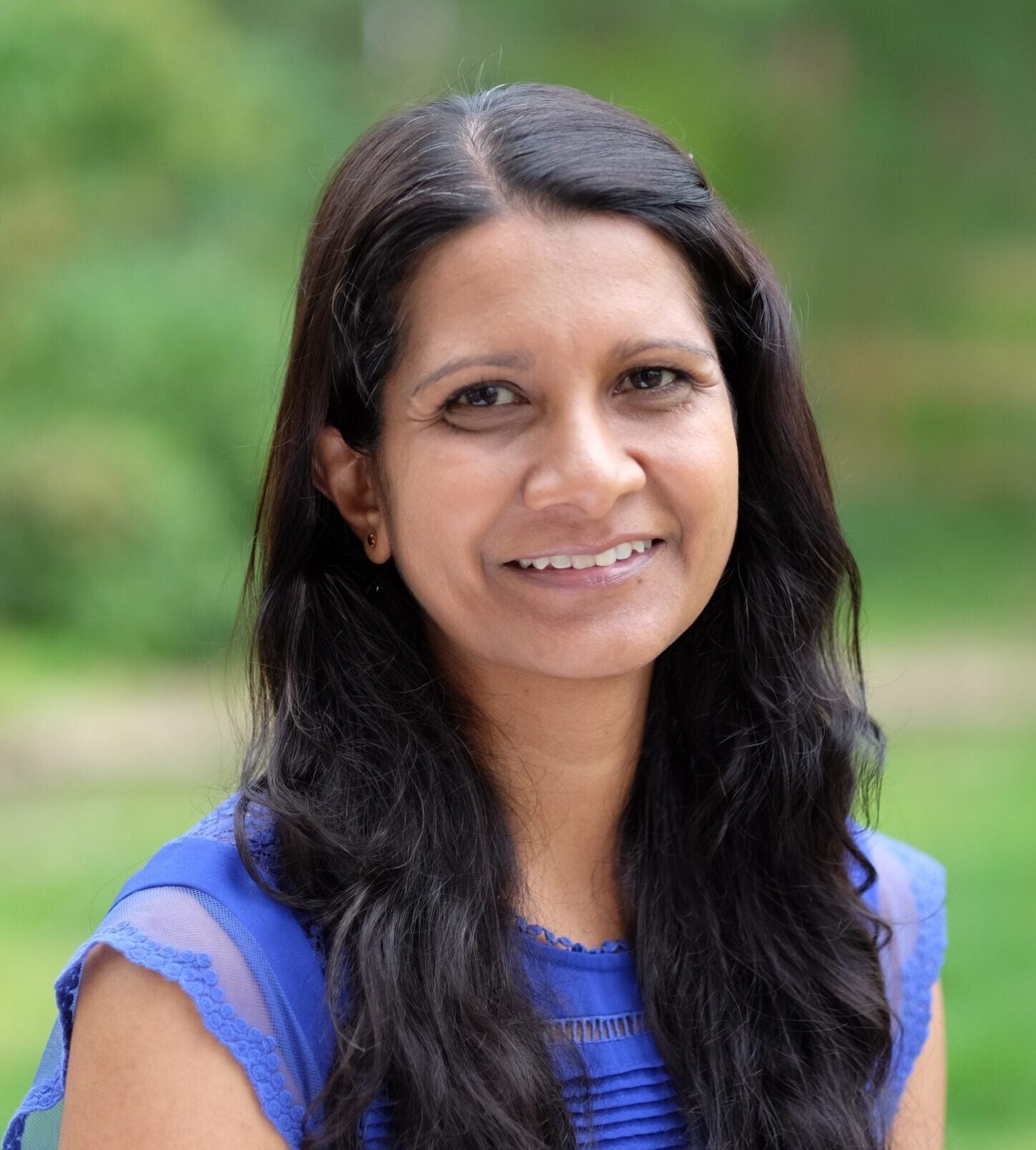 Dr. Sudhir smiling with blurred green background and wearing blue sleeveless blouse