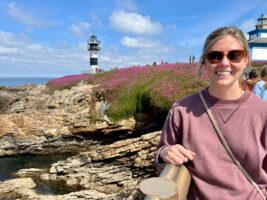 Gabriella smiling outside and in front of a rocky terrain with flowers and a lighthouse