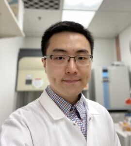 Michael Chi smiles while wearing a lab coat and glasses