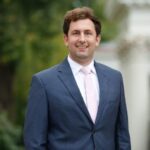 Clint Upchurch wearing a suit smiles and stands in front of greenery