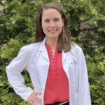 UVA Pharmacology Graduate Student Blair Towers stands in front of greenery smiling and wearing a pink shirt and white lab coat.