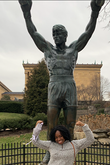 University of Virginia Prisca Obidike, MD, Surgery Resident raises her arms to celebrate near a statue with raised arms.